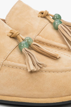 A close-up shot of the front of the shoe, displaying the artisanal decorative tassel with a green bead and the soft, matte texture of the light suede.