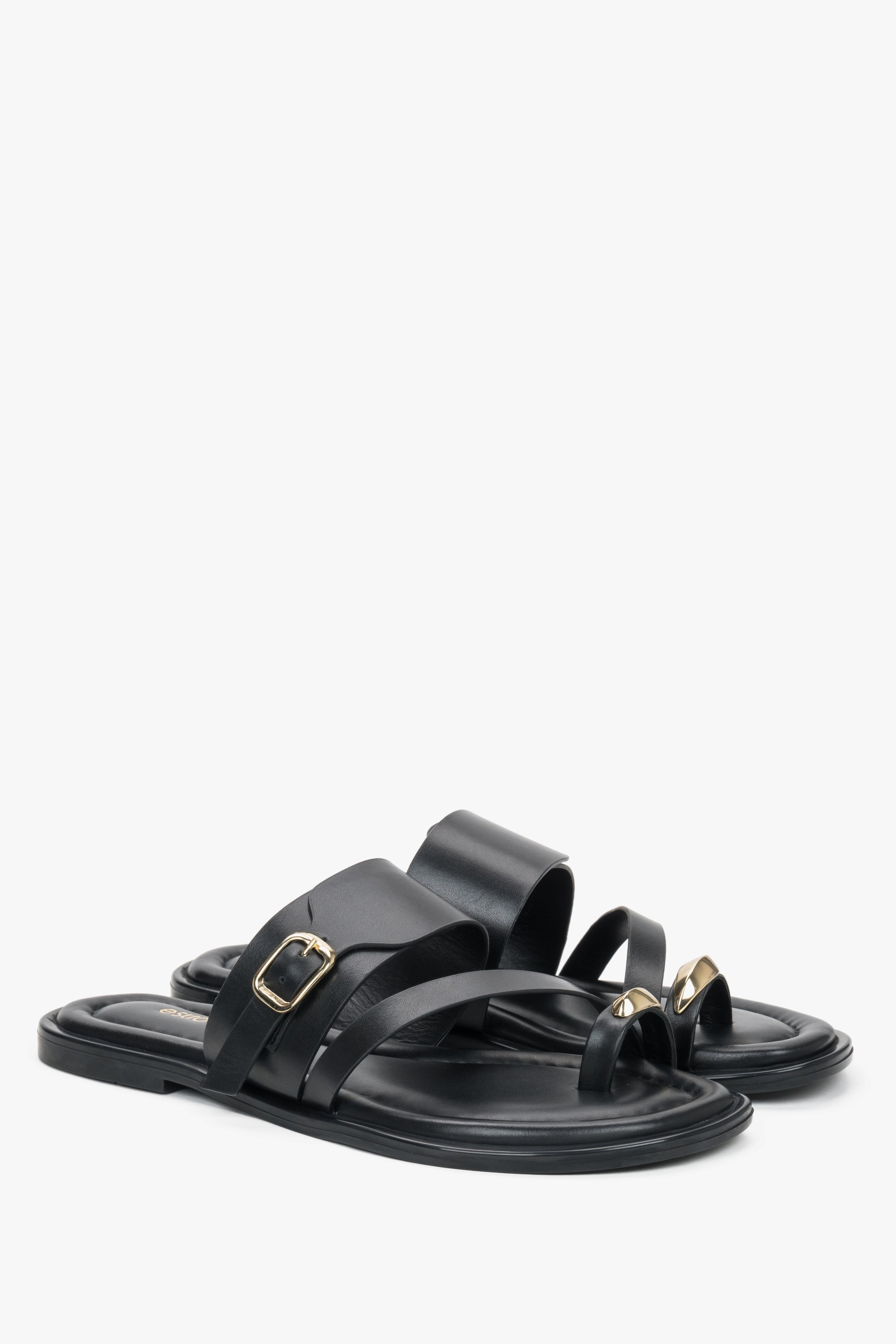 Black leather Estro mules with a toe loop, showcasing an asymmetrical upper line and a bold gold buckle detail.