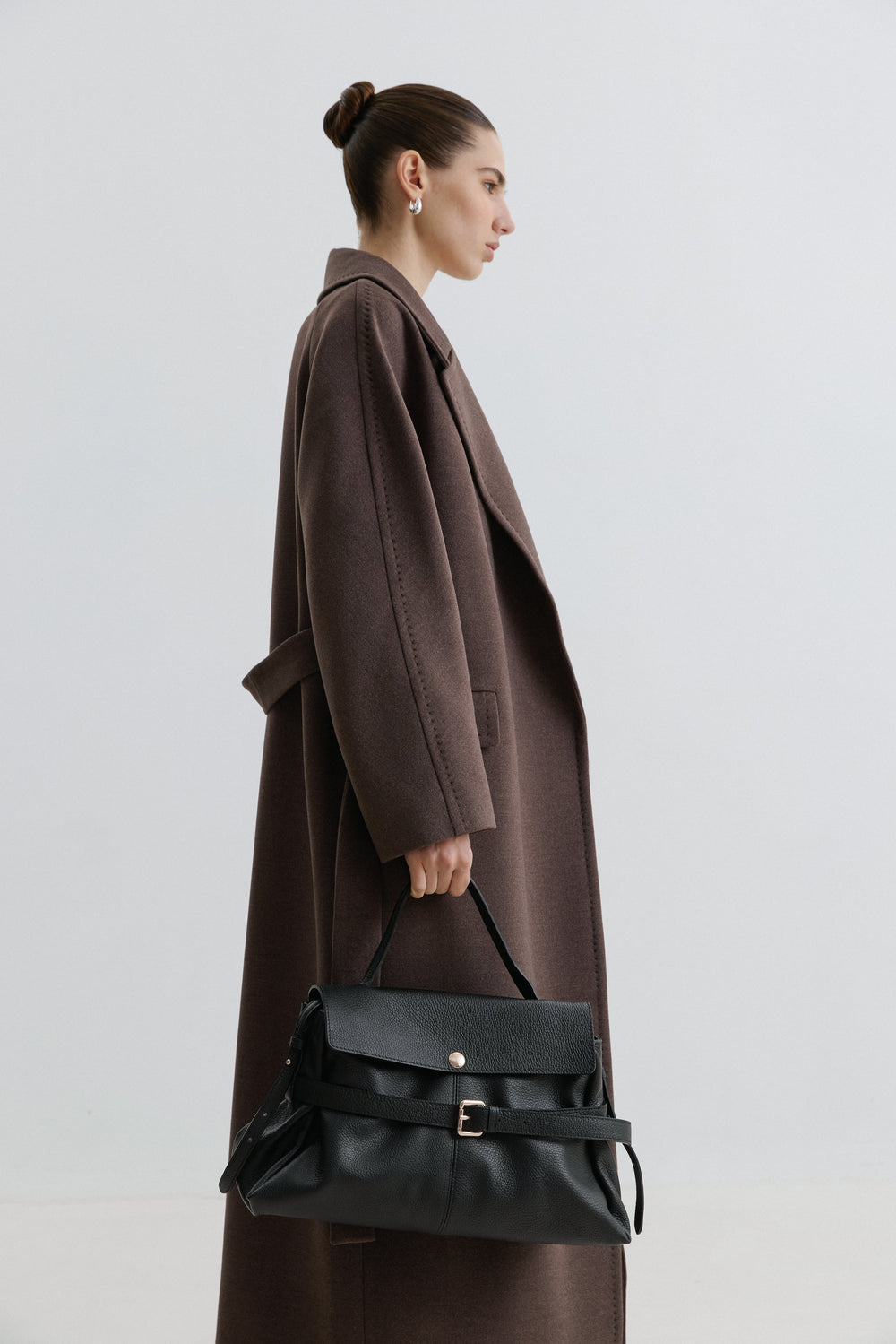 Woman wearing a long brown coat holding a black handbag against a plain background.