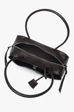 Elongated dark brown shoulder bag. Compact natural leather silhouette with a decorative bag charm.