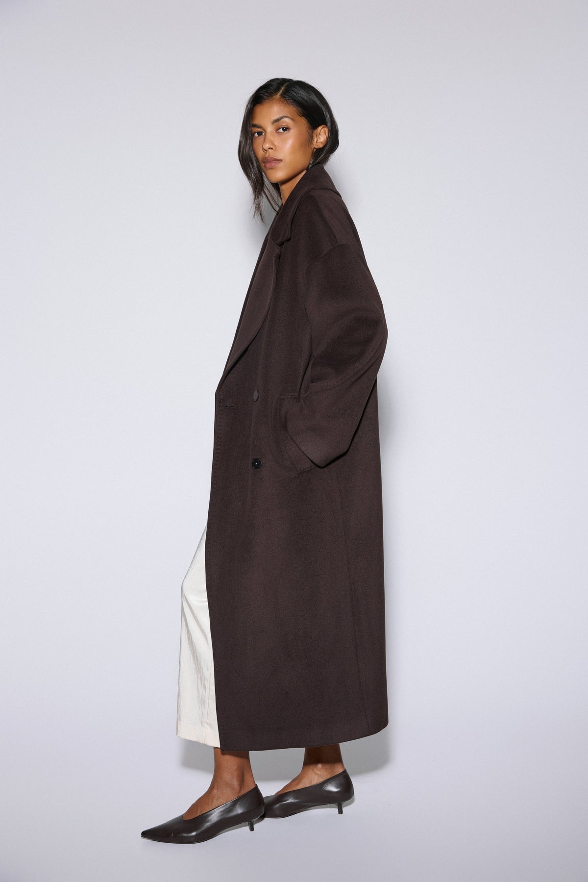 Women's long dark brown wool coat Estro - side profile view displaying the fluid drape and tie belt accent.
